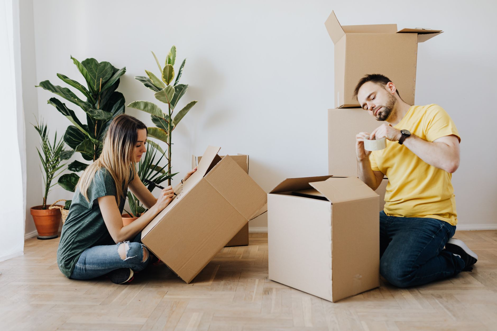 Side view full length glad couple packing stuff into cardboard boxes while sitting on floor at home during relocation in new