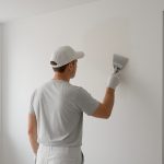 An individual in a gray shirt and gloves painting a wall with a spatula in a bright room.